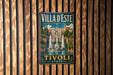 A vintage-style metal sign with the text "Villa d'Este" and "Tivoli Italy" displayed on it, hanging on a wooden wall.