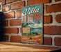 A vintage-style metal sign for Villa Louis Wisconsin, featuring a scenic image of a lake and a boat, mounted on a brick wall.