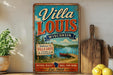 A vintage-style metal sign for Villa Louis Wisconsin, featuring a scenic lake and surrounding landscape, is displayed on a shelf next to a wooden cutting board and some potted plants.
