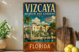 A vintage-style metal sign for the Vizcaya Museum and Gardens in Florida, featuring a tropical landscape with palm trees and a fountain.