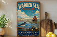 A vintage-style poster of the Wadden Sea in Netherlands, featuring a lighthouse, a sailboat, and various marine life, is displayed on a wall next to a wooden cutting board and a potted plant.