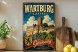 A vintage-style metal sign displaying the name "Wartburg Castle" and the word "GERMANY" in bold letters, with a castle illustration and a coat of arms. The sign is placed on a wooden surface, with a potted plant, a wooden cutting board, and some lemons nearby.