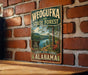 A vintage-style metal sign for Weoguffee State Forest in Alabama, featuring a scenic landscape with trees, a lake, and mountains.