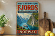 A vintage-style sign advertising West Norse Fjords, featuring a scenic image of a waterfall and a ship, is displayed on a shelf next to a potted plant and a wooden cutting board.