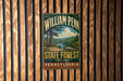 A vintage-style metal sign for William Penn State Forest in Pennsylvania, featuring a scenic landscape with pine trees and a lake.