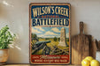 A vintage-style metal sign for Wilson's Creek National Battlefield in Missouri, featuring a cannon and a monument, is displayed on a shelf next to a wooden cutting board and some lemons.