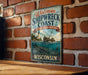 A vintage-style metal sign for the Wisconsin Shipwreck Coast, featuring a lighthouse and a shipwreck, is displayed on a brick wall.