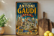 A vintage-style metal sign with the text "WORKS OF ANTONI GAUDI SPAN" and an illustration of the Sagrada Familia, a famous Barcelona landmark. The sign is displayed on a shelf next to a wooden cutting board and a potted plant.