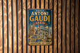 A vintage metal sign with the text "WORKS OF ANTONI GAUDI SPAIN ARCHITECTURE ART VISION" is displayed on a wooden wall.