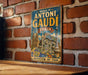 A vintage metal sign with the text "WORKS OF ANTONI GAUDI SPAN" and an illustration of the Sagrada Familia, a famous Barcelona landmark, is displayed on a brick wall.