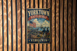 A vintage metal sign for Yorktown Battlefield in Virginia, featuring an American flag, cannons, and a ship, mounted on a wooden wall.