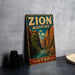 A vintage-style metal sign with the text "ZION NARROWS UTAH" is displayed on a gray concrete surface, with a stack of black camera lenses and a white dish visible to the side.