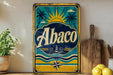 A vintage-style metal sign with the word "Abaco" prominently displayed, featuring a sun, palm trees, and a lighthouse, is placed on a shelf next to a wooden cutting board and some lemons.