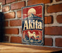 A metal sign with the word "Akita" on it, featuring a dog and a mountain landscape, is mounted on a brick wall.