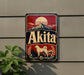 A vintage-style metal sign with the word "Akita" prominently displayed, featuring a mountain landscape and a dog.