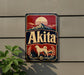 A vintage-style metal sign with the word "Akita" prominently displayed, featuring a mountain landscape and a dog.