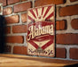 A vintage-style metal sign with the word "Alaska" prominently displayed, mounted on a brick wall.