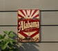 A vintage-style sign with the word "Alaska" prominently displayed, featuring a sunburst design and a small building. The sign is mounted on a gray wall, and there is a plant visible to the left of the sign.