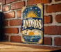 A vintage-style metal sign with the word "ANDROS" displayed prominently, featuring a lighthouse and a sun-filled sky.