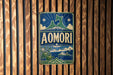 A vintage-style metal sign with the word "AOMORI" displayed prominently in the center, surrounded by various decorative elements such as stars, mountains, and a ship.