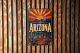 A vintage-style metal sign with the word "ARIZONA" prominently displayed, featuring a desert landscape with mountains and a sunburst design.