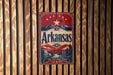 A vintage-style metal sign with the text "Arkansas" and a landscape illustration, mounted on a wooden wall.