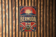 A vintage metal sign with the word "BERMuda" prominently displayed, featuring a sun, palm trees, and a lighthouse, all set against a wooden background.