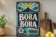 A vintage-style metal sign with the text "BORA BORA" displayed prominently, featuring a tropical landscape with palm trees, a mountain, and a sun. The sign is placed on a shelf next to a wooden cutting board and a potted plant.