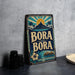 A vintage-style metal sign with the text "Bora Bora" is displayed on a gray countertop, with a stack of black camera lenses and a white dish visible to the side.