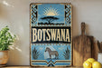 A vintage-style metal sign with the word "Botswana" prominently displayed, featuring a zebra and other wildlife elements. The sign is placed on a wooden surface, with a potted plant, a wooden cutting board, and some lemons nearby.