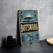 A vintage-style metal sign with the word "Botswana" prominently displayed, featuring a zebra and other wildlife elements. The sign is placed on a gray surface, with a stack of black camera lenses and a white dish visible in the background.