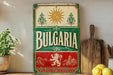 a vintage-style sign with the word "Bulgaria" prominently displayed, surrounded by decorative elements such as a sun and a lion. The sign is placed on a wooden surface, with a potted plant, a cutting board, and some lemons nearby.