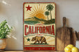A vintage-style metal sign with a bear and palm trees, along with the word "California" prominently displayed, is placed on a wooden surface.