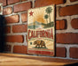 A vintage-style metal sign with the word "CALIFORNIA" prominently displayed, featuring a sun, palm trees, and a bear.