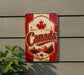 A vintage-style metal sign with the word "Canada" prominently displayed, featuring a maple leaf and a scenic mountain landscape.