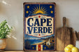 a vintage-style metal sign with a blue background, red and white border, and a sun and star design. The sign prominently displays the text "CAPE VERDE" in large letters. The sign is placed on a shelf next to a wooden cutting board and a potted plant, creating a rustic and charming atmosphere.