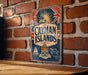 A vintage-style metal sign with the text "CAYMAN ISLANDS" and various symbols, including a sun, palm trees, and a ship, is displayed on a brick wall.