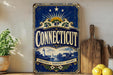 A vintage-style metal sign with the word "Connecticut" prominently displayed, featuring a lighthouse and other scenic elements. The sign is placed on a wooden surface, with a potted plant and a wooden cutting board nearby.