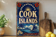 A vintage-style sign for the Cook Islands, featuring a tropical island scene with a sailboat, palm trees, and a sun. The sign is displayed on a shelf next to a wooden cutting board and a potted plant.