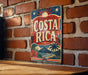 A vintage-style metal sign with the text "COSTA RICA" displayed prominently, featuring a tropical landscape with palm trees and mountains.