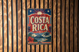 A vintage-style metal sign with the text "COSTA RICA" displayed prominently in the center, surrounded by tropical imagery such as palm trees, flowers, and a sunburst design.