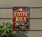A vintage-style metal sign with the text "COSTA RICA" displayed prominently, featuring a tropical landscape with palm trees, a mountain, and a sun.