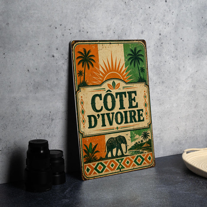 A vintage-style metal sign with the text "COTE D'IVOIRE" displayed prominently, featuring an elephant and palm trees.