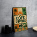 A vintage-style metal sign with the text "COTE D'IVOIRE" displayed prominently, featuring an elephant and palm trees.