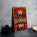 A vintage-style metal sign with the word "DENMARK" prominently displayed, featuring a cityscape and a sailboat illustration. The sign is placed on a gray surface, with some black objects, such as a stack of cups, visible in the background.