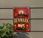 A vintage-style metal sign with the word "DENMARK" prominently displayed, featuring a cityscape and a sunburst design.