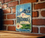 A vintage-style metal sign with a blue background and a red star, featuring the word "Djibouti" and a depiction of a coastal town.