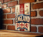 A vintage-style sign with the word "ENGLAND" prominently displayed, featuring a red and white color scheme with a crown and a lion emblem. The sign is mounted on a brick wall, and there is a wooden table below it.