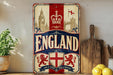 A vintage-style metal sign with the word "ENGLAND" prominently displayed, featuring a red and white color scheme with a crown and a lion emblem. The sign is placed on a wooden surface, with a potted plant and a wooden cutting board visible in the background.