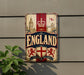 a sign with the word "ENGLAND" prominently displayed, along with a red and white shield and lion emblem. The sign is mounted on a wall, and there is a plant visible to the left of the sign.
